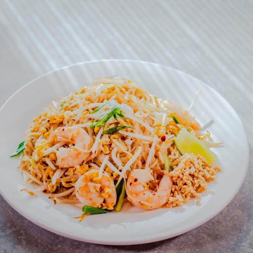 A white plate of shrimp pad thai with bean sprouts, lime, and crushed peanuts.
