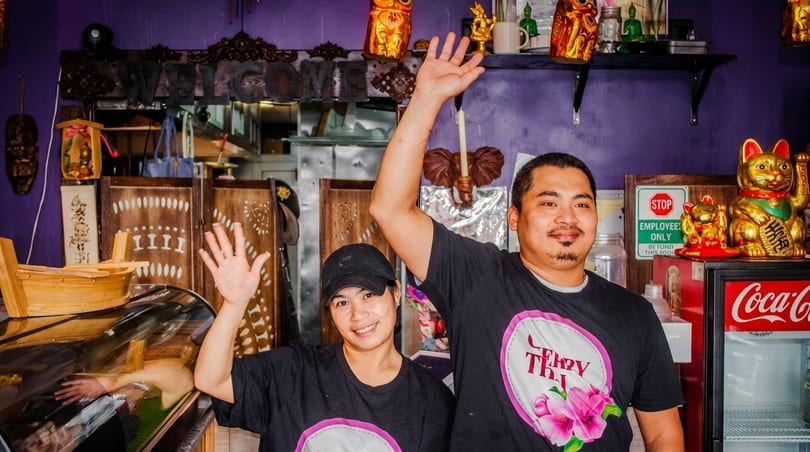 Two smiling restaurant staff members in black t-shirts wave from behind a counter with Thai decor.