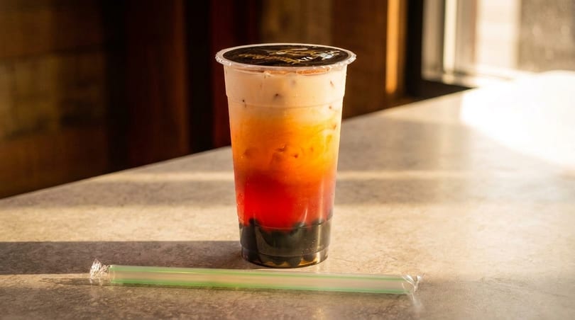Layered bubble tea with red, orange, and white colors, and dark boba pearls, on a sunlit counter.