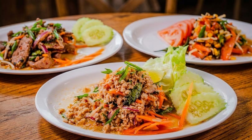 Three plates of Thai food, including larb, beef salad, and papaya salad, on a wooden table.