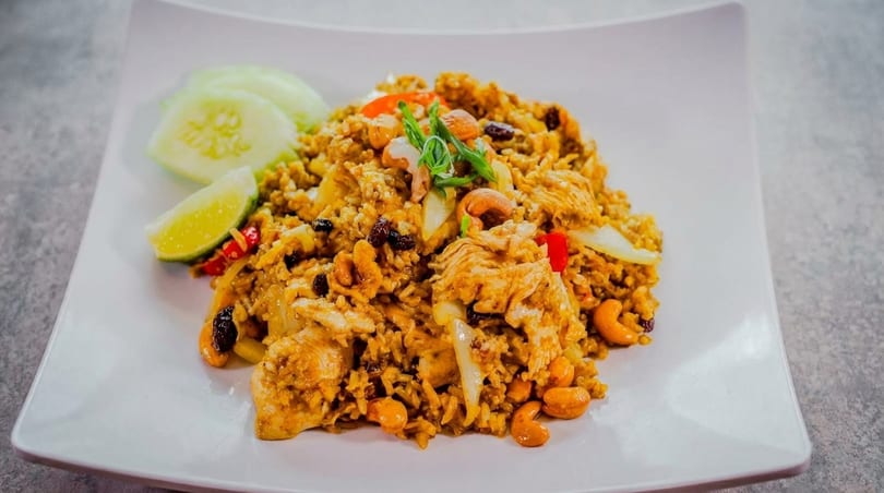 Chicken fried rice with cashews, raisins, and vegetables, garnished with lime and cucumber slices.