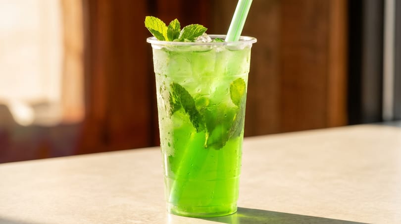 Bright green drink with ice and fresh mint leaves in a clear plastic cup with a straw.